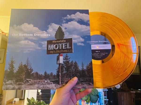 A hand holds a transparent orange vinyl record as it pokes out of its sleeve, which depicts the sign of burned down motel that reads: Fireside Motel Restaurant.
