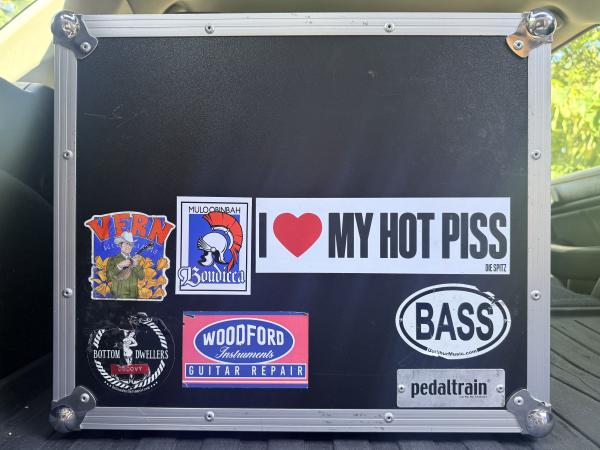 Guitar pedalboard case with band stickers on it, prominently featuring an I Love My Hot Piss Die Spitz bumper sticker.